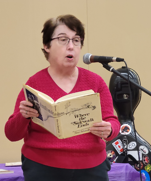 A woman standing at a microphone reading from a book titled "Where the Sidewalk Ends"