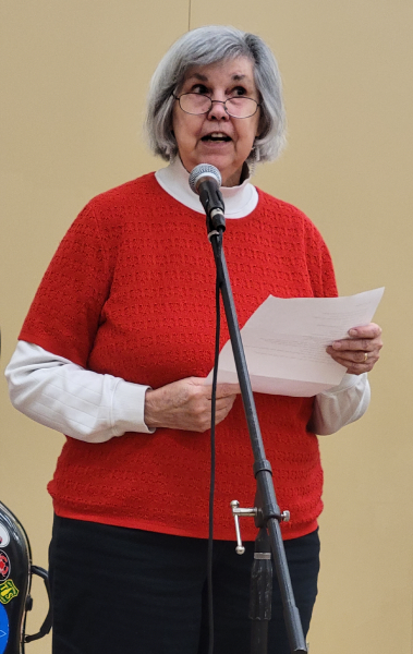 A woman standing at a microphone reading a piece of paper being held